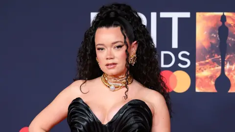 Getty Images A woman wearing a black dress, and sparkling gold and silver jewellery around her neck. She is standing in front of a board which reads "Brit Awards".