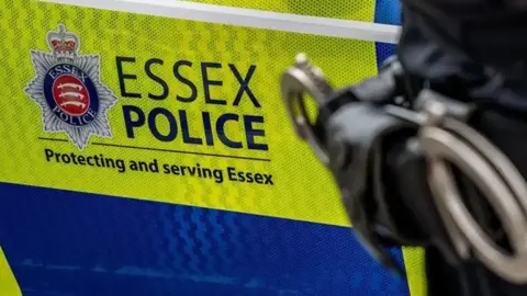 The side of a police car that is fluroesent yellow and blue, and a sign that says ESSEX POLICE with the forces' logo. An officer is standing next the car with handcuffs on their left pocket.