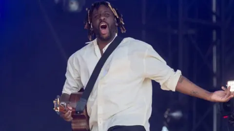 Myles Smith, wearing a white shirt and black trousers, sings during Radio 1's Big Weekend in Sunderland, England in May. He has a guitar strapped across his shoulder 