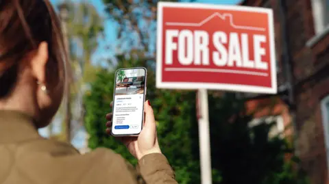 Woman looks at smart phone screen about a property with a for sale sign in the background.