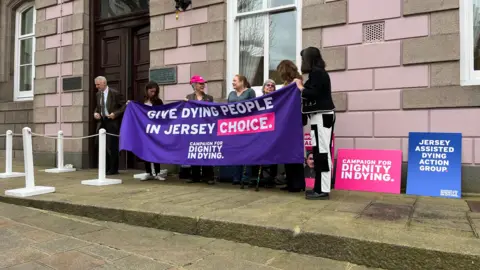 A group of people stand outside a building holding a banner that reads "give dying people in Jersey a choice".