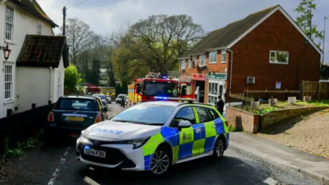 Colin Jackson A police car is parked in the middle of the road and fire engines can be seen in the background. Homes and small businesses are either side of the road.