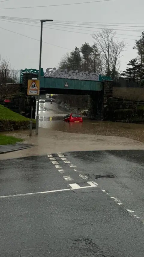A junction on a rainy day. Flood water can be seen pooling under a bridge and there is a small red car in the middle of it.