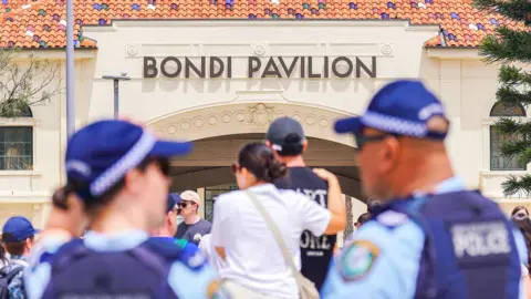 Anadolu via Getty Images Police officers are seen in the foreground with mourners in the background in front of the Bondi Pavilion on 15 December.