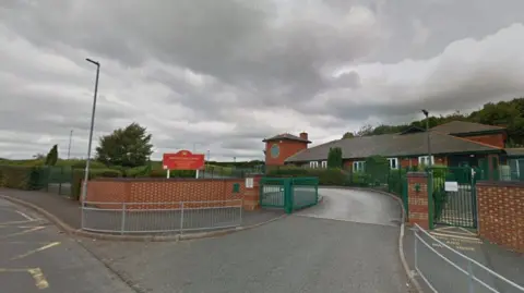 The entrance to the car park of Goldenhill Primary Acdemy, with a red sign indicating the name of the school. There is a single-storey building in the background with a number of bushes and trees around.