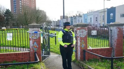 PA Media A policewoman stands at the entrance to Rawnsley Park, near the scene in the St Philips area of ​​Bristol where a 16-year-old boy died after being stabbed. There is police tape opposite the door, and colorful houses and a tall tower block can be seen in the background.