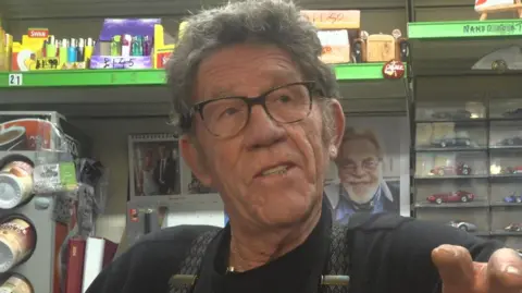 A grey haired man, with glasses, wearing a black t-shirt and braces