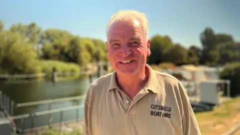A man with light hair stood in front of the river wearing a shirt which reads: "Cotswold Boat Hire"