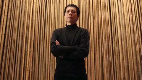 Getty Images a man dressed all in black, and with short black hair, stands in front of a wall of wooden poles