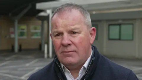 Steve McManus is wearing a white buttoned shirt, with the top button undone, and a blue zipped-up jumper outside the hospital.