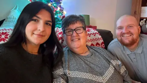 Picture shows three people sat on a sofa smiling. Woman on far left of picture is wearing a black top and has long black hair. She is sat next to a woman on the right who is wearing a grey top and glasses and has short grey hair. She is sat next to a man who is on the far right of the picture who is wearing a grey top and has a bald head and a ginger beard and moustache.