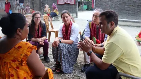 JOA Aid workers from Jersey Overseas Aid talk to a woman in an orange dress with red flowers on in an outdoor area.