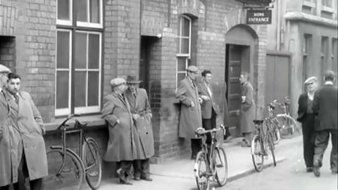 Black and white image of a number of men in long coats and hats standing outside the jobs and benefits office.  A sign for 'Mens Entrance' is above one of the doors.  A number of bicycles are sitting around.