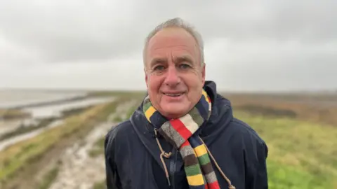 Shaun Whitmore/BBC Man with short white hair wearing a scarf with coloured stripes and a raincoat