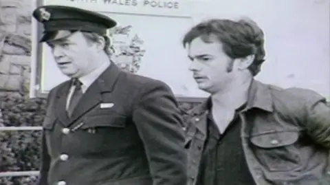 Black-and-white picture of a prisoner (on the right) being escorted by a prison officer. The prisoner is wearing a denim jacket and has a moustache.