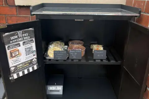Steph Goodhand The inside of a cake shed - it is painted black and has baked goods inside with chalkboard signs. She has a locked payment box and posters advertising how to pay.