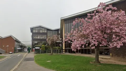 The outside of a school in Scarborough, with a road leading to the school buildings. To the right of the image is a green space with cherry blossom trees in full bloom. 
