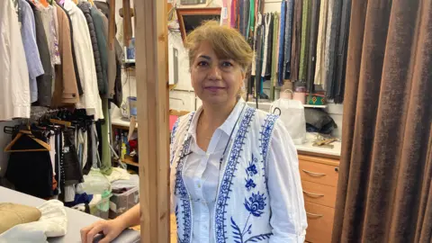 A woman in a white blouse and blue-and-white patterned waistcoat stands in a tailors stall at an indoor market. 