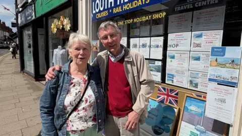 Carol Steels and husband David, both in their 60s, are standing in front of a travel agents in Lincoln's high street