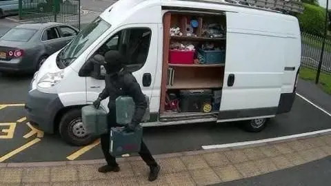 @TheGasExpert A man in a black coat carrying three toolboxes away from a white van with its doors open.