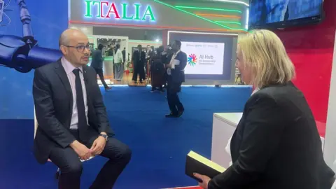 Sir Demis Hassabis, wearing a dark suit and tie, speaking to Zoe Kleinman at the summit.