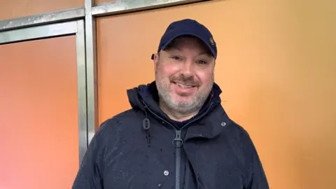 A white male wearing a dark coloured rain coat and baseball cap stares at the camera