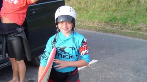 Supplied A young Laura Crane smiles at the camera as she holds a surf board in one arm. She is wearing a white helmet and a blue Oneill rashvest over a black wetsuit. 