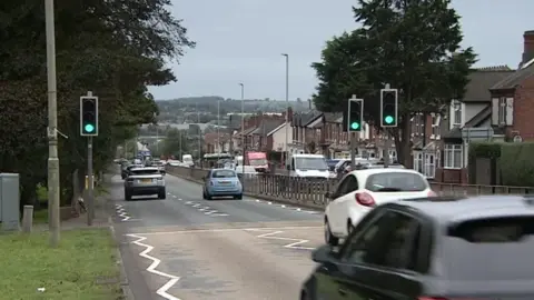 BBC A view of a road with cars driving down two lanes. Houses line the right of the road and grass and trees are on the left hand side of the road.