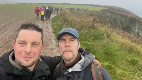 Maxwell Family Fund Craig Maxwell smiles and has an arm around comedian Rhod Gilbert's shoulder. The pair are in the middle of a muddy path with a group of hikers in the background.