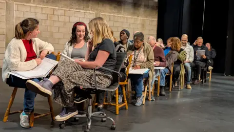 Kevin Middleton A group of actors set on wooden chairs in a long line. The line has two chairs in each line and the actors are reading scripts. In the front three woman are turned to each other to talk. 