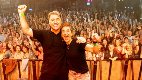 Day Fever  Jonny Owen and Vicky McClure with their arm around each other, the opposite arms aloft, as they stand on stage with a crowd of people cheering and waving behind them 