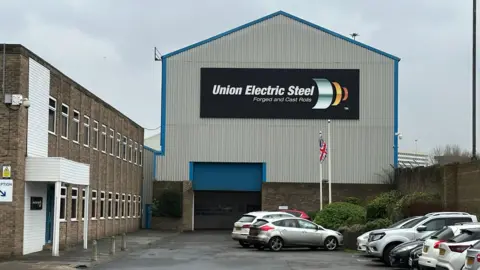 A front-on view of the Union Electric Steel building. There are parked cars in front of the blue entrance. A brick building sits to the left side.