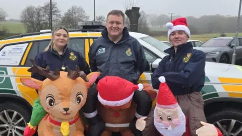 One woman and two men wearing dark jackets and in inflatables costumes, looking like they are the backs of a reindeer, gingerbread man and a Santa. The people all standing in front of a yellow and white car