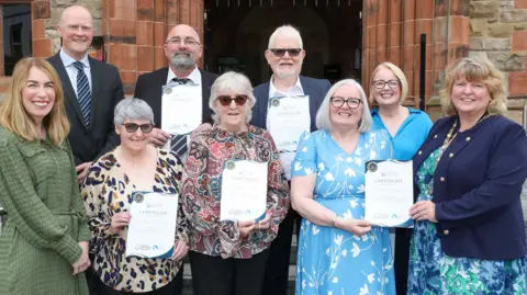 Tourism NI Three men and six women stand should to shoulder in two rows outside the red brick building of Derry's Guildhall. They are being presented with certificated.