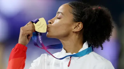 Jazmin Sawyers is sideways on to the camera as she kisses her medal, in her right hand. She has tied-back black frizzy hair and is wearing a white team top.