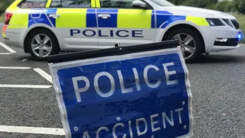 BBC Police sign reading "police accident". Behind the sign is a white police vehicle with yellow and blue check patterning.