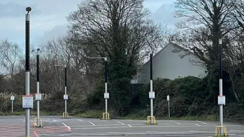 Conflow Power Group A car park in which iLamps are visible - the lamps have dark cylindrical solar cells at the top and a yellow metal frame protects the base