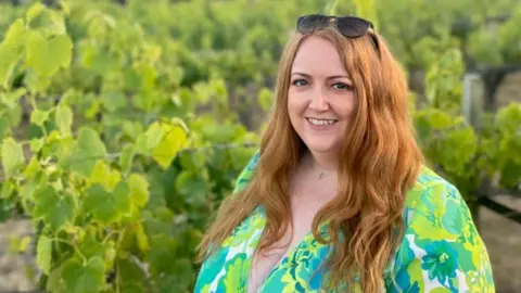 Family Handout Moyra Whelan wearing a green floral patterned dress and standing in a vineyard. She is smiling at the camera and has long red hair with sunglasses resting on her head. The background is slightly blurry and it looks like a warm summer evening.