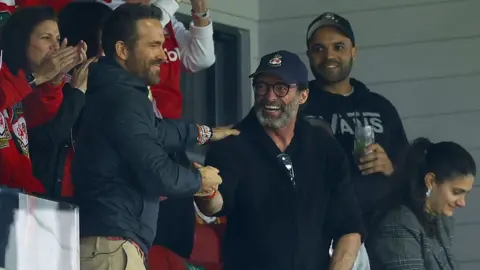 Getty Images Ryan Reynolds shakes the hand of Hugh Jackman from the stands at the Racecourse Ground