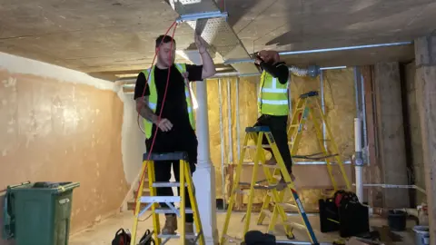 Two men in high visibility vests stood on yellow step ladders. They are working on a fixture which comes down from the ceiling. There are bare walls, a green bin and some tools in the background.