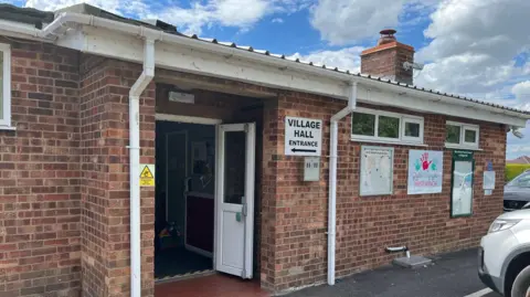 Clare Worden/BBC West Winch village hall is a one storey brick building. On the outside are lots of signs including "Village hall entrance" with an arrow pointing to an open door. 