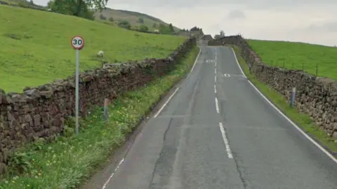 A country road runs between two fields, one of which has a sheep in. There is a 30mph sign at the side of the road.