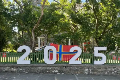 A 2025 handmade sign to show support for the 2025 island games, with orkney flags in the background 