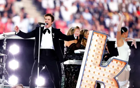 PA Media Brandon Flowers performs with The Killers at the UEFA Champions League final. He's dressed in a tuxedo with a bow tie, standing next to a large billboard-style letter K.