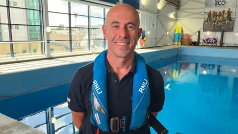 A man with an RNLI life jacket on stood in front of an indoor swimming pool.