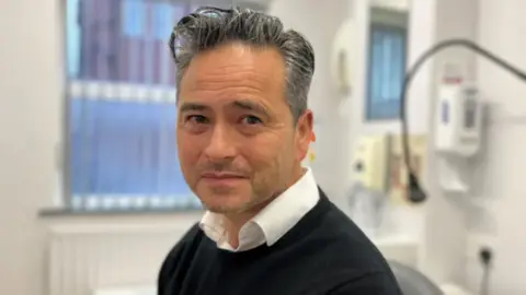 A middle-aged man with combed grey hair, wearing a shirt and jumper, looks towards the camera in a medical office, and is pictured in medium close up.