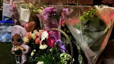 Several bunches of flowers with cards attached lined up on the pavement against a brick wall. There are also two cuddly toys, one of a doll-like figure on a purple cushion and a second of a blue Stitch character in a ziplock bag.
