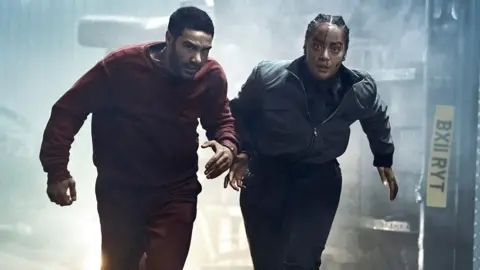 Rahim (left) and Hoyle (right) in a still from new series Prisoner. They are pictured running towards the camera while handcuffed together. Rahim wears a dark red sweatshirt and tracksuit trousers. Hoyle wears black trousers, a grey bomber jacket, shirt and tie. She has blood on her face.