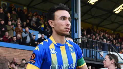 Michael Clunan is standing on a football pitch near a stand. He has short dark hair and is wearing a blue and yellow King's Lynn Town-shirt which has branding on it. 