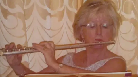 An older woman with mid-length blonde hair and glasses looks down as she plays the flute and reads sheet music. She is wearing a sleeveless glittery silver dress and is wearing a dainty watch on one wrist and a bracelet on the other. She is stood against a ruffled gold and white curtain. 